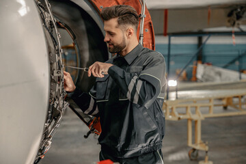 Smiling man airline maintenance technician using screwdriver while repairing airplane at repair station