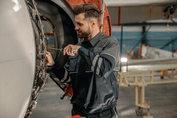 Smiling man airline maintenance technician using screwdriver while repairing airplane at repair...