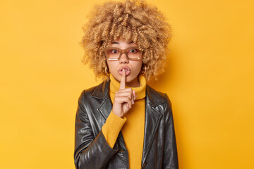 Mysterious curly haired woman makes silence gesture presses index fingers to lips asks to be quiet makes taboo sign wears sweater and leather jacket isolated over yellow background. Do not speak - obrazy, fototapety, plakaty