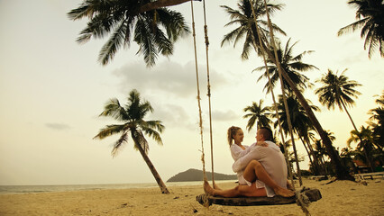 Cute loving couple on the swing on tropical beach. Woman and man in white shirt enjoy gold sunset and life in beaches, palm tree. Evening warm sunset. Summer holiday vacation tropical tourism concept