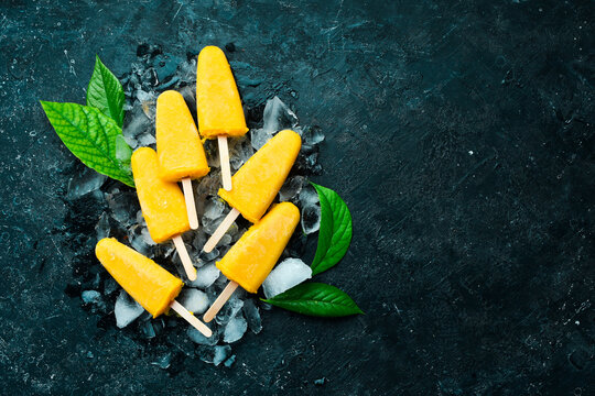 Frozen Yellow Popsicles On Ice. Cold Summer Fruit Sweets. On A Black Stone Background. Top View.