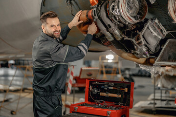 Cheerful man maintenance technician looking at camera and smiling while repairing airplane at...