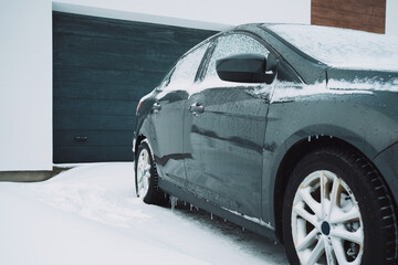 a gray car is covered with snow against the background of a garage of a private house