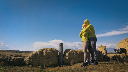 Man and woman in yellow green sportswear. Lovely couple of travelers hug and kiss near old stone enjoying highland landscape. Two travelers are walking against the backdrop of snow-capped mountains.