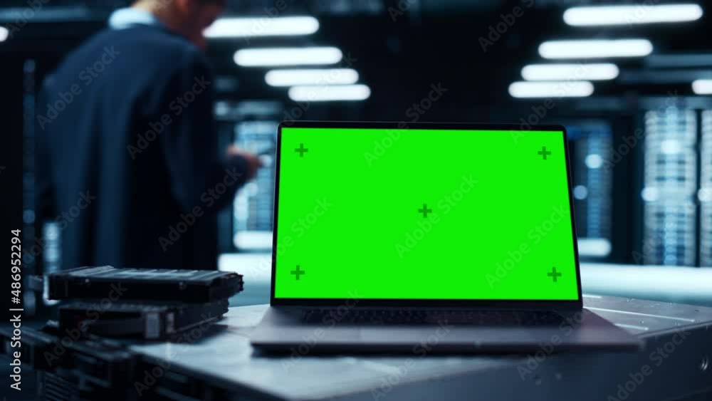 Poster Laptop Computer with Green Screen Chroma Key Mock Up Display Stands on a Table. In the Background Data Center With Rows of Server Racks and IT Engineer Working. Static Shot.
