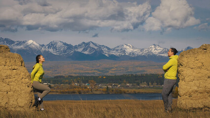 Man and woman in yellow green sportswear. Lovely couple of travelers hug and kiss near old stone enjoying highland landscape. Two travelers are walking against the backdrop of snow-capped mountains.