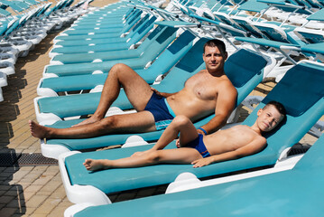 A young athletic man and his son are smiling happily and sunbathing on a sun lounger on a sunny day at the hotel. Happy family vacation at a hotel in the resort. Summer holidays and tourism.