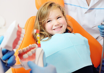 pediatric dentist shows how to properly brush her teeth to a little girl who is sitting in an orange dental chair and smiling.
