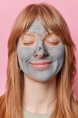Close up shot of pleased teenage girl stands with eyes closed applies mud mask takes care of complexion and skin has long straight red hair isolated over pink background. Facial treatment concept