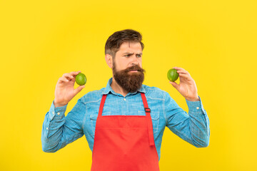 Worried man in red apron looking at fresh limes citrus fruits yellow background, fruiterer