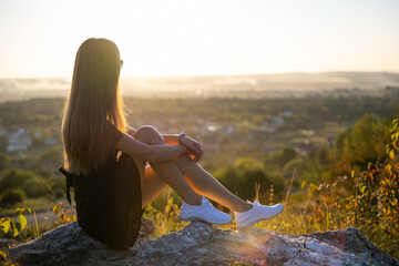 Young elegant woman in black short dress and white sneaker shoes sitting on a rock relaxing outdoors at summer evening. Fashionable lady enjoying warm sunset in nature