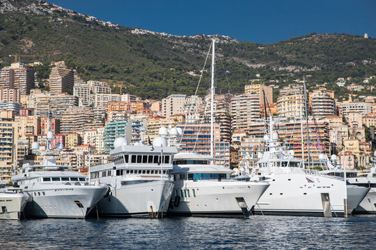 A lot of huge yachts are in port of Monaco at sunny day, megayachts are moored in marina, is a yacht show, Monte Carlo, real estate housing is on background, glossy board of the motor boat