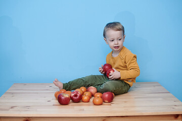 Fototapeta premium 2 year old boy with red apples and tangerines. boy dressed green pants yellow jacket sits advertising vegetables fruits