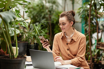 Young smiling businesswoman in brown blouse scrolling in mobile phone while communicating or...