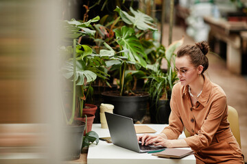 Young contemporary female designer or owner of small business working in the internet while sitting by desk in front of laptop in florist shop - obrazy, fototapety, plakaty