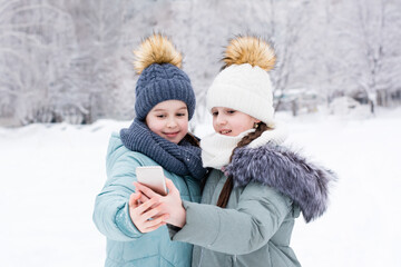 Fototapeta na wymiar Two smiling girls in warm clothes take a selfie on a smartphone in a snowy winter park. Lifestyle use of technology