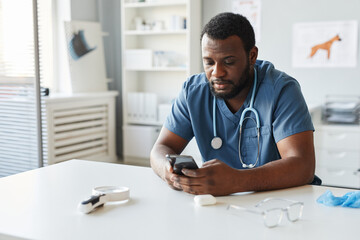 Young African American veterinarian in blue uniform scrolling in mobile phone while sitting by workplace in hospital - obrazy, fototapety, plakaty