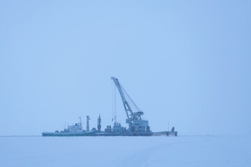 Fototapeta premium Floating crane for loading and unloading and installation work moored in winter in a frozen bay. Snowfall.