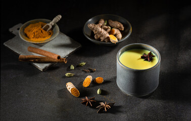 A glass cup of golden milk with turmeric powder and fresh turmeric root and anise and cinnamon on black. The concept of spices and herbs. Ayurvedic drink on a light background. Space for text.