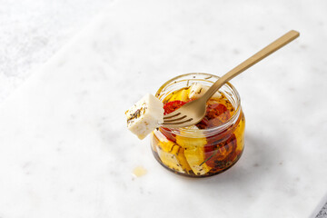 Fork with organic feta cheese in olive oil in glass jar with tomatoes. Product on marble background