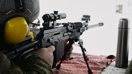 A sniper in hearing protection aims at a target at the firing line, a thermos with warm tea is...