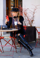 Side view of busy female blogger, which wearing in vogue apparel, hat on head and leather boots, using smartphone for work and holding cup of coffee while sitting outside cafe