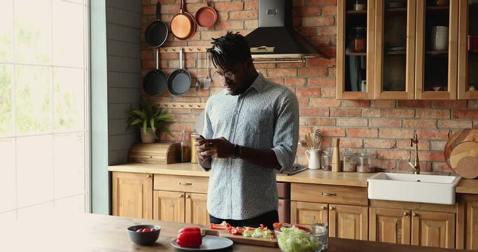 Young African man stand by kitchen table use phone learn to cook at online master class surf recipe at culinary website. Millennial husband text message to wife ask advice how to prepare tasty salad - Powered by Adobe