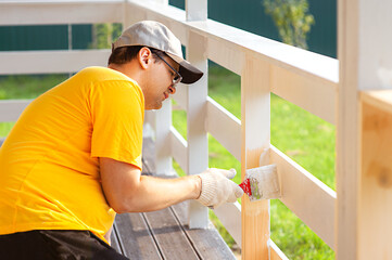 A man in a bright yellow T-shirt and cap holds a brush in his hand and paints the boards on the porch of the house with white paint