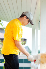 A man in a bright yellow T-shirt and cap holds a brush in his hand and paints the boards on the porch of the house with white paint