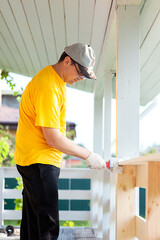 A man in a bright yellow T-shirt and cap holds a brush in his hand and paints the boards on the porch of the house with white paint