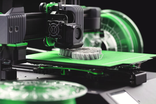 oblique view on FDM-3D-printer with printed parts while manufacturing white helical gears. brightly lit scenery with green build plate and spare green filament on transparent rolls