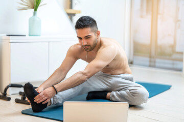 Young ordinary man, beginner stretching with tow hands to one leg while watching online workout class on laptop at home.
