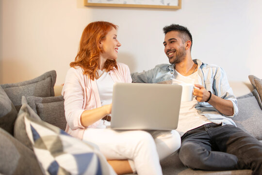 Happy friends having relaxed conversation while using laptop and sitting on sofa indoors.