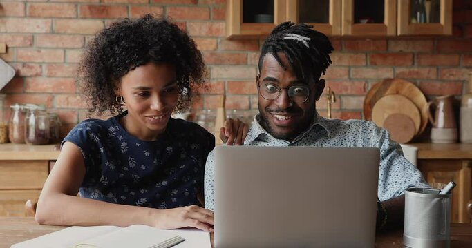 Overjoyed African family couple look on laptop screen at kitchen laugh give high five getting great online offer mortgage approval in official email. Excited young spouses become web lottery winners