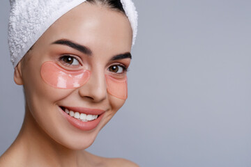 Beautiful young woman with under eye patches and hair wrapped in towel on grey background, space for text