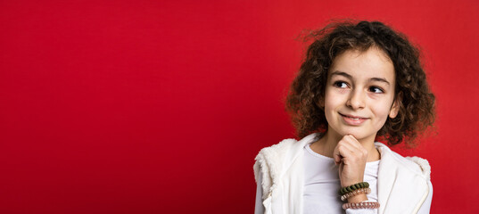 One small caucasian girl ten years old with curly hair front view portrait close up standing in front of red background looking to the camera smiling hand on chin thinking idea copy space