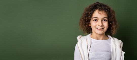 One small caucasian girl ten years old with curly hair front view portrait close up standing in front of green background looking to the camera smiling happy and joy copy space