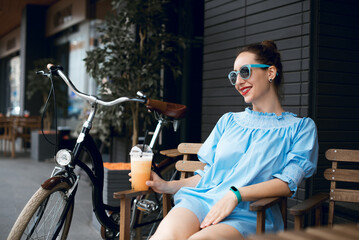 Fototapeta na wymiar Young beautiful lady drinking fresh smoothies on a sunny summer day during her ride. Riding a bicycle in the city. Bicycle tourism. Refreshing fruit drink. Active rest. Hipster on a bike