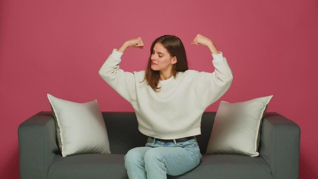Proud young woman raising arms showing biceps demonstrate hands muscles, satisfied with achievements, sitting on sofa
