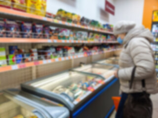 Fototapeta na wymiar Defocus woman holding supermarket basket blurred background with different products and standing near refrigerator. Supermarket aisle colorful shelves of merchandise. Retail concept. Out of focus