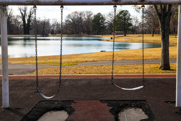 a rusty white metal swing set near a still green lake in a park surrounded by yellow winter grass,...