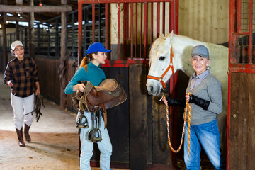 Women ranchers preparing white horse for ride. Senior woman leading horse out of stable, younger woman carrying saddle, Asian woman carrying tack.