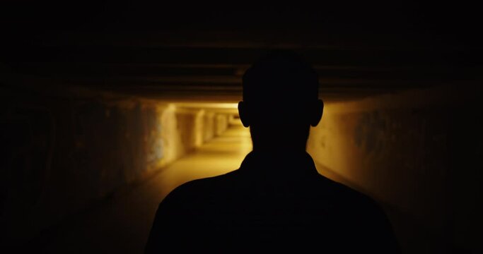 Silhouette of the back of a man walking in a dark underpass. Dark scary movie scene at night city. Handheld camera shot