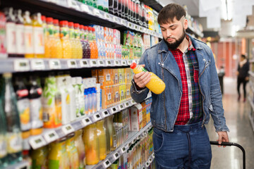 Young glad cheerful positive bearded man choosing bottled fresh juices in grocery section at...