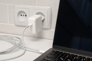 Charging laptop with power adapter in electrical socket on white table, closeup