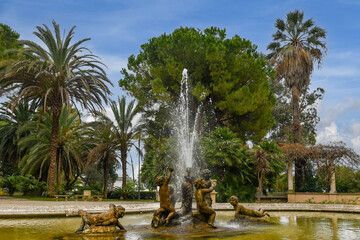 The stone fountain with putti or cherubs in the gardens of Villa Ormond (1889) with palms and maritime pines in the background, Sanremo, Imperia, Liguria, Italy - obrazy, fototapety, plakaty
