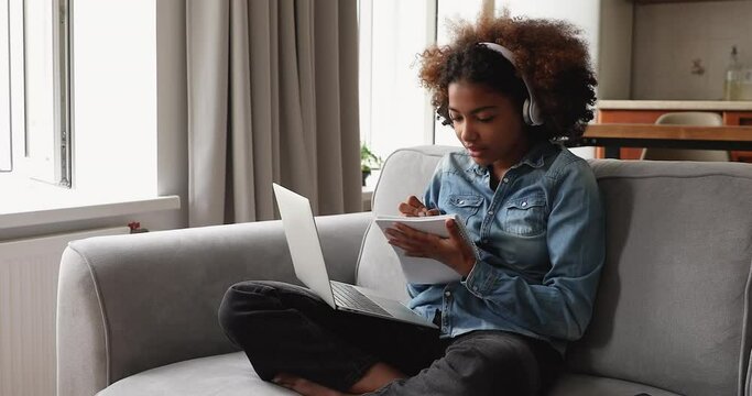 Cute 18s African student girl sit on sofa wear headphones take notes while studying remotely use laptop and video call app. Gen Z and modern tech usage, distancing learning at home, tuition concept
