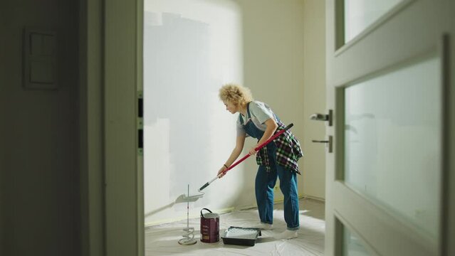 Single woman is painting wall in gray in a empty room using paint rollers. Door in foreground