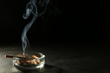 Smoldering cigarette in glass ashtray on grey table against black background. Space for text