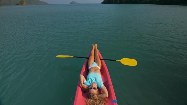 Pretty slim woman in elegant swimsuit lies in pink plastic kayak drifting on azure ocean at exotic resort upper view. Traveling to tropical countries. Girl is sailing on kayak in ocean, view above.